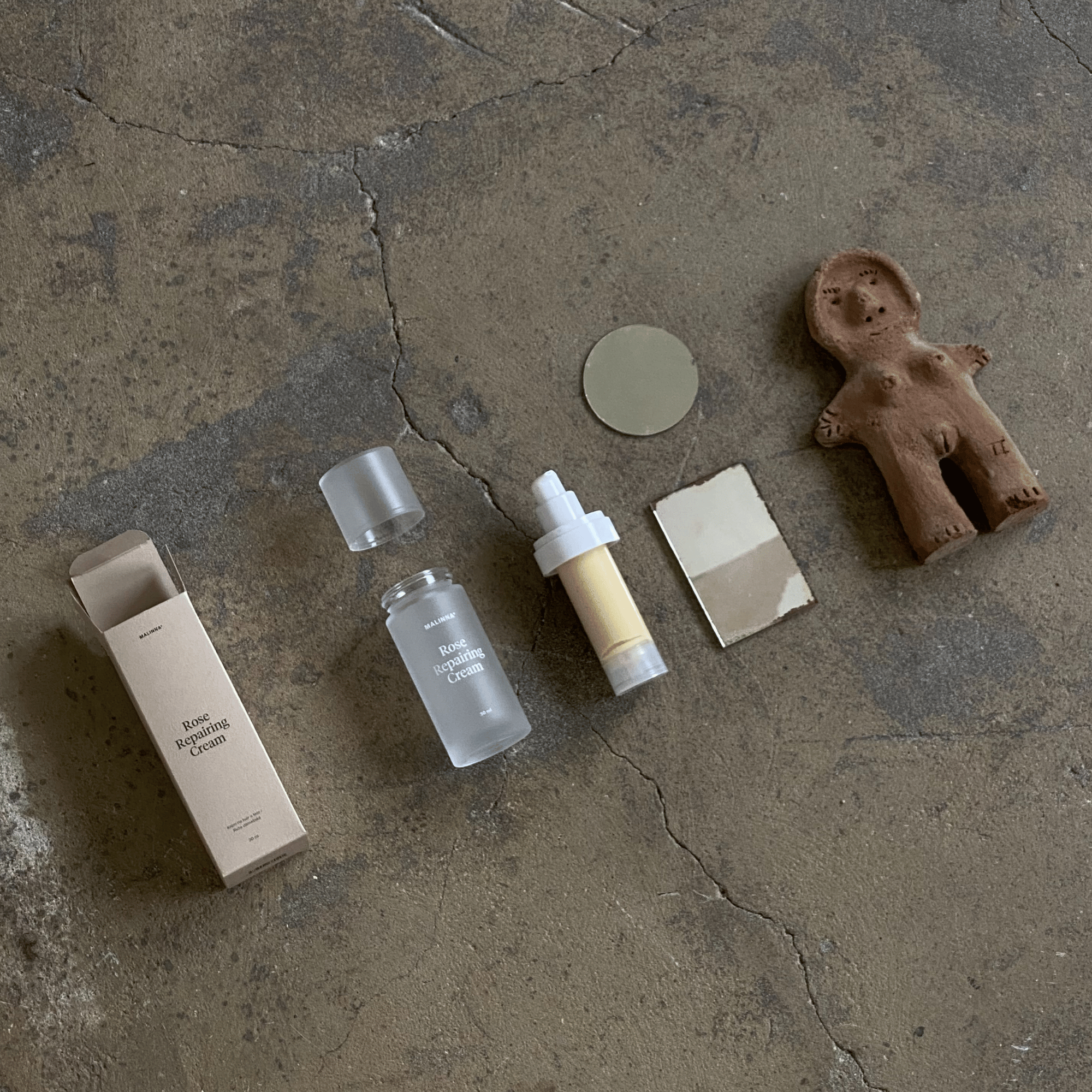MALINNA Rose Repairing Cream rozložený a položený na kamennej zemi vedľa zrkadielok a hlinenej sošky