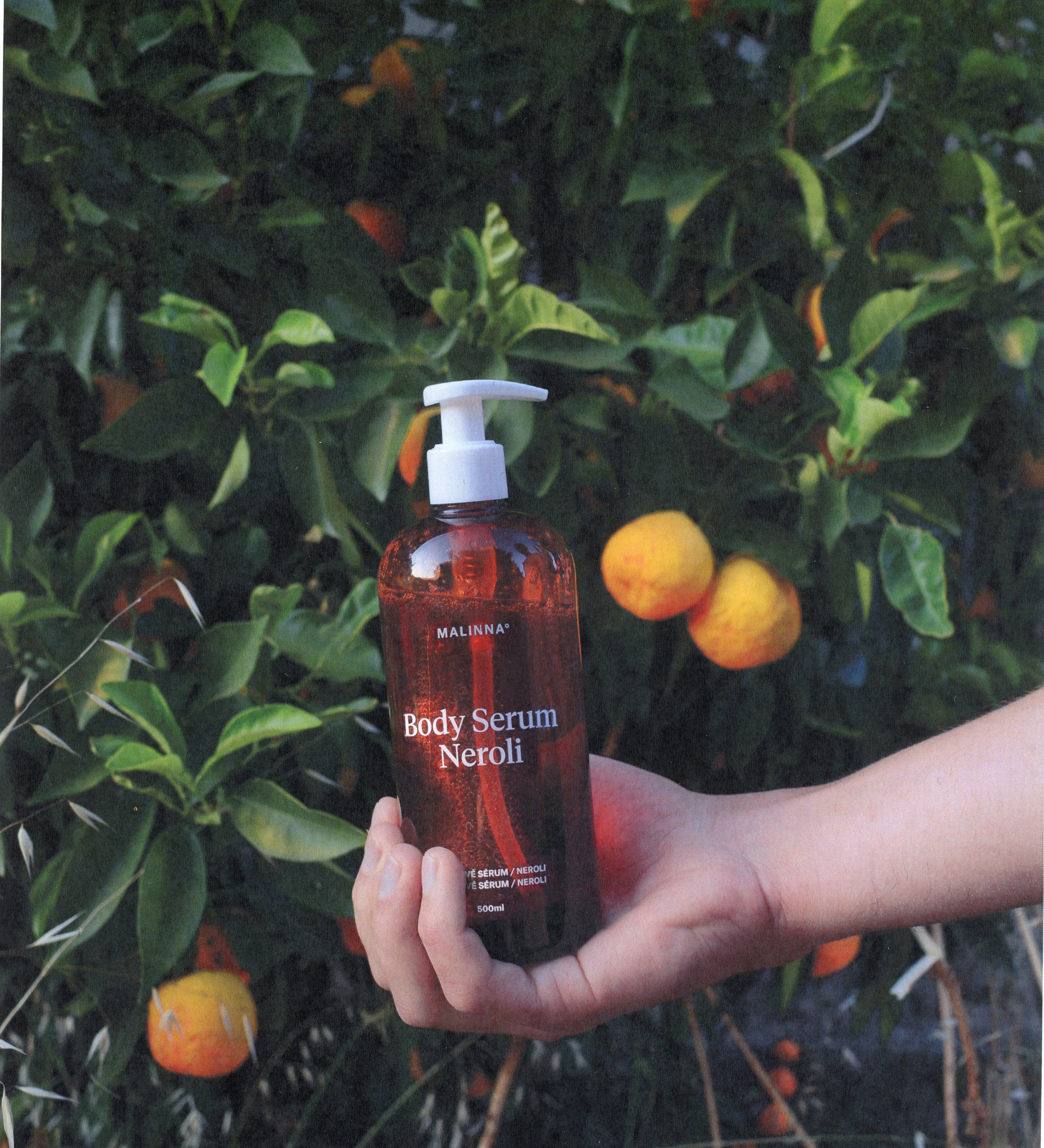 Ruka držiaca MALINNA Body Serum Neroli pri strome pomarančovníka