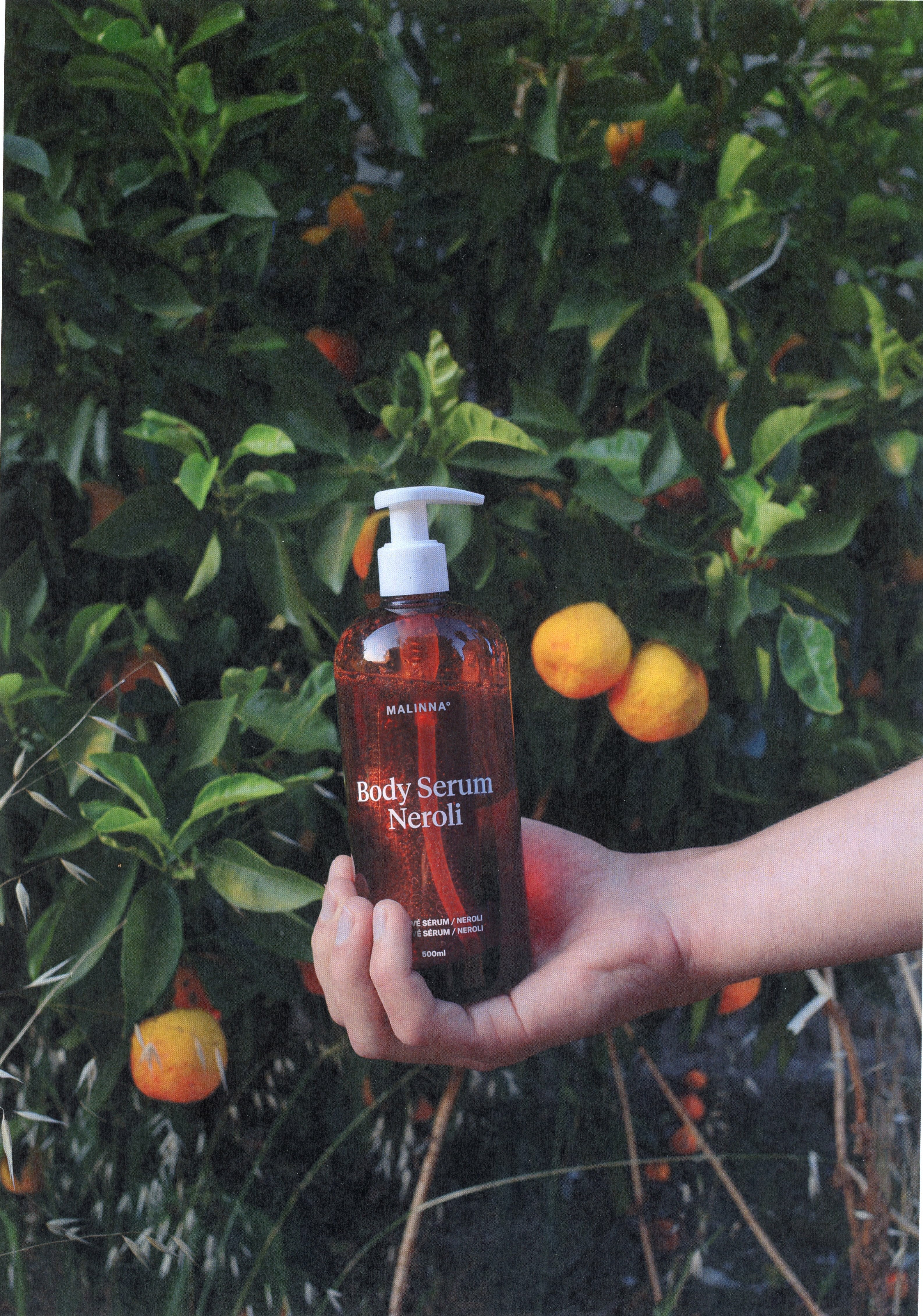 Ruka držiaca MALINNA Body Serum Neroli pri strome pomarančovníka